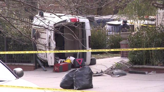 A white SUV lays on its side behind police tape. Part of a gate has been broken and items have slipped out of the vehicle's trunk. 