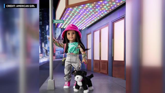 An American Girl doll is posed next to a toy streetlight with a "Broadway" street sign and a toy dog on a leash. 