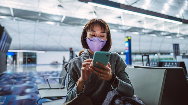 Young Asian woman in protective face mask using smartphone joyfully while waiting for night flight in airport terminal 