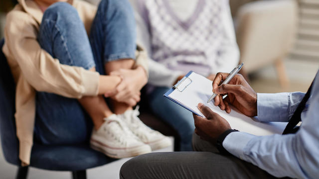 Man holds a clipboard in front of two teens on a couch 