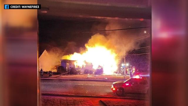 Flames engulf a one-story home while firefighters stand outside. 