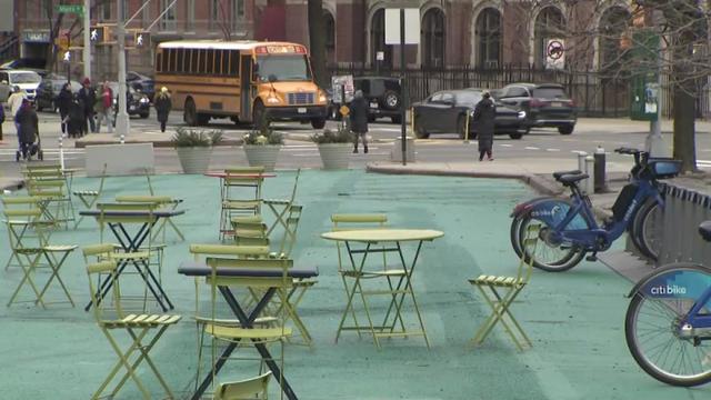 Empty tables and chairs sit in a pedestrian plaza near a bike rack with Citi Bikes. 
