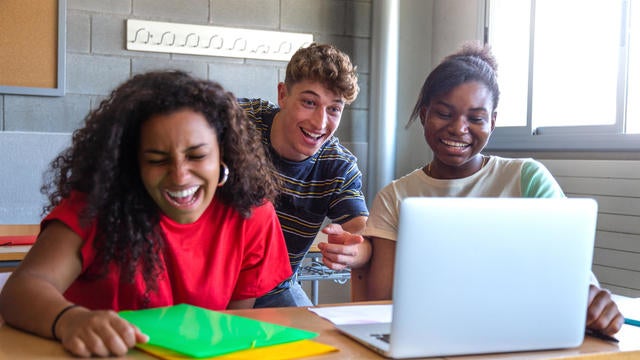 Multiracial group of high school students laughing together in class using laptop to do homework together. 