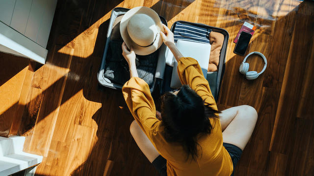 Overhead view of young Asian woman sitting on the floor in her bedroom, packing a suitcase for a trip. Getting ready for a vacation. Traveller's accessories. Travel and vacation concept 