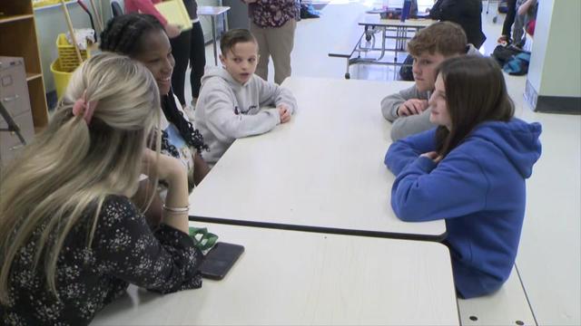 Four middle school students sit at a cafeteria table with a teacher. 