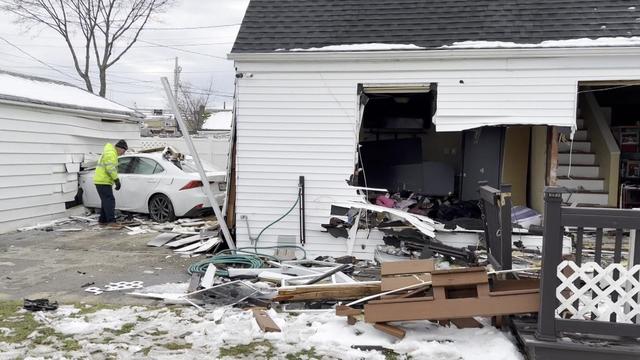 A large hole in seen in the front of a single-story house. In the background, a vehicle can be seen crashed into the side of another building that appears to be a shed or garage. 