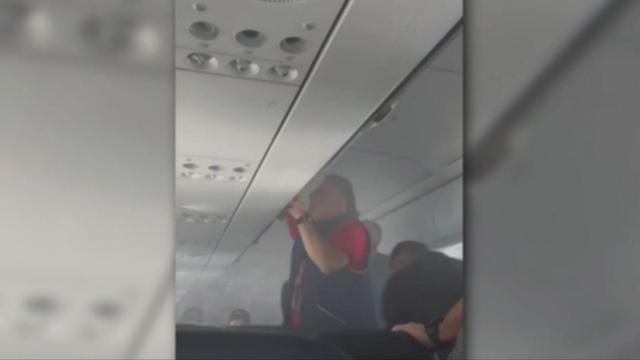 A retired firefighter stands in the aisle of a plane and looks in an overhead bin. 