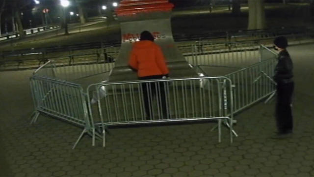 One person spray-paints a Christopher Columbus statue in Central Park while another person watches. 