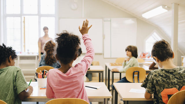 Rear view of boy raising hand while answering in class at elementary school 