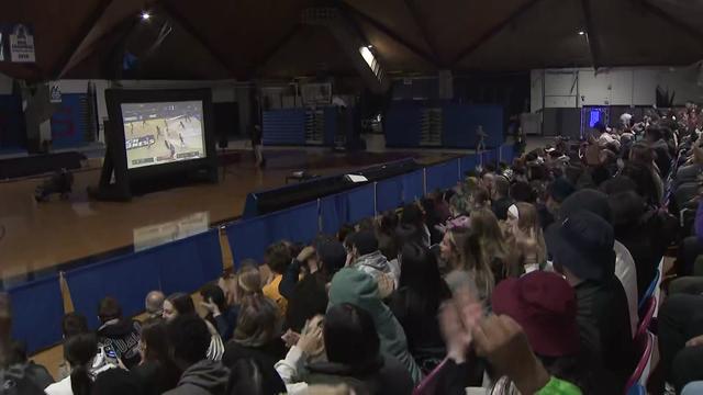 A crowd sits on auditorium bleachers watching a televised basketball game on a large screen. 