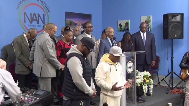 Rev. Al Sharpton stands behind a podium at the National Action Network surrounded by a group of people bowing their heads in prayer. 