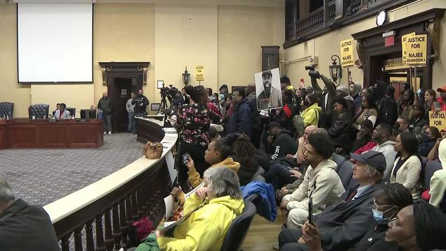 Community members sit at a Paterson City Council meeting, some holding signs reading "Justice for Najee Seabrooks." 
