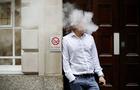 White smoke surrounds the head of a man leaning against a wall with "no smoking" sign 