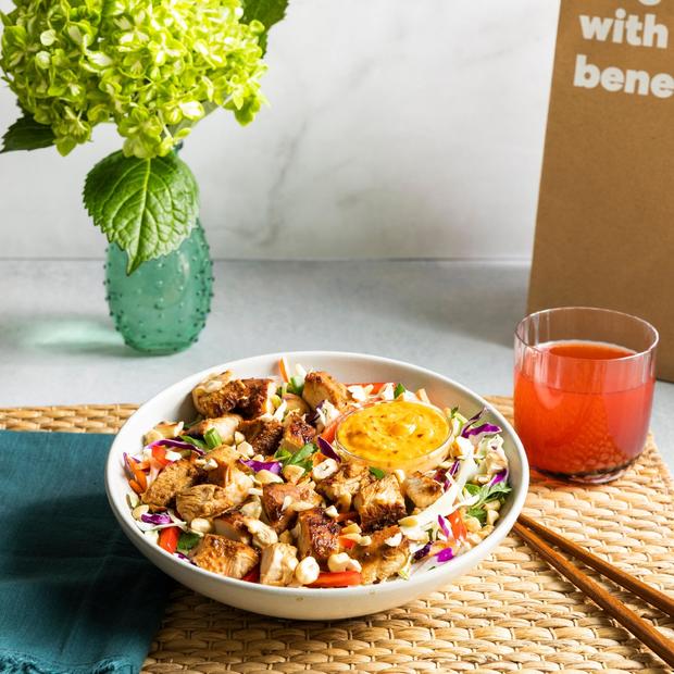 Asian Chopped Salad in a bowl next to chopsticks, light orange beverage, a flower in a green decorative vase, and a delivery bag 