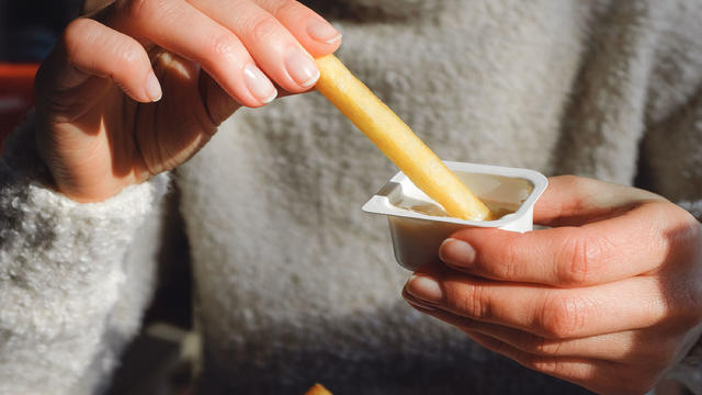 A woman holds French fries in her hand, eating them in a cafe or restaurant. A girl in a white knit sweater dips a slice of potato in garlic sauce. The concept of fast food, snacking, junk food. 