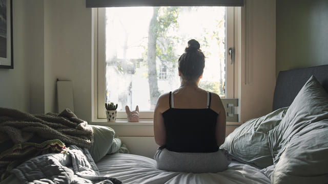 Rear view of a teenage girl looking out of the window whilst sitting on her bed 