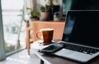 Cropped shot of wooden coffee table with laptop, smartphone and a cup of tea in the living room at home by the window against beautiful sunlight 