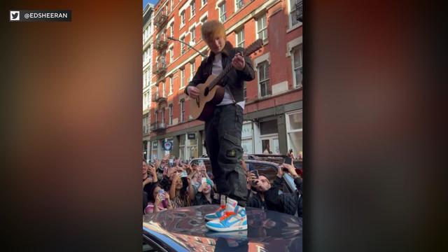 Ed Sheeran stands on top of a parked car on a New York City street, playing an acoustic guitar. Dozens of fans surround the car, many holding up smartphones to photograph or record Sheeran. 