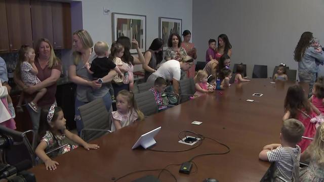 A number of women and small children stand and sit around a long conference table. 