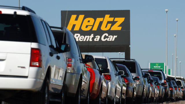 A row of rental cars are parked below a sign for Hertz Corp. 