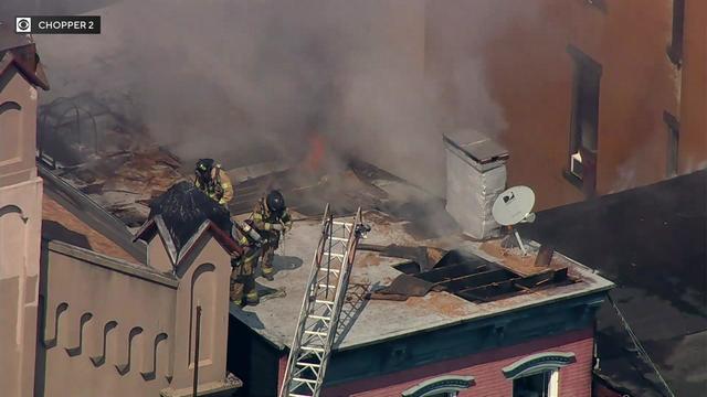 Several firefighters stand on the roof of a building. Flames can be seen coming through the roof. 