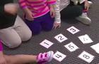 Young children kneel on the ground near flashcards with numbers on them. 