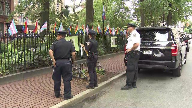 Three NYPD members stand on a sidewalk looking at a display of Pride flags on an iron fence. 