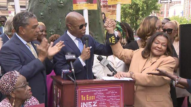 Mayor Eric Adams stands behind a podium raising the hand of Assemblymember Inez Dickens. 