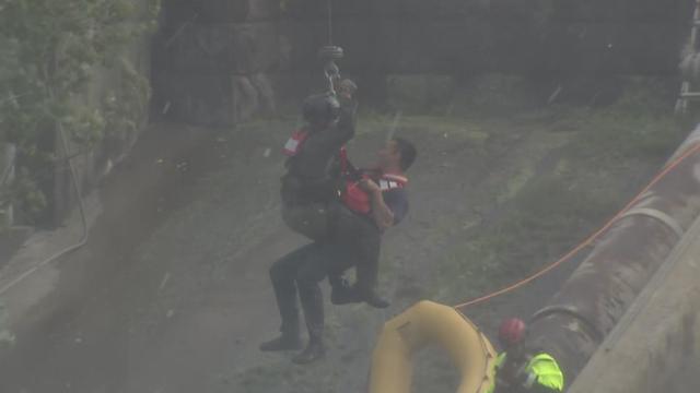 Two people on a harness being hoisted into a helicopter over the Passaic River. 