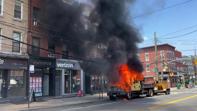 Flames engulf the front of a Department of Transportation truck parked on a street in front of several stores. 