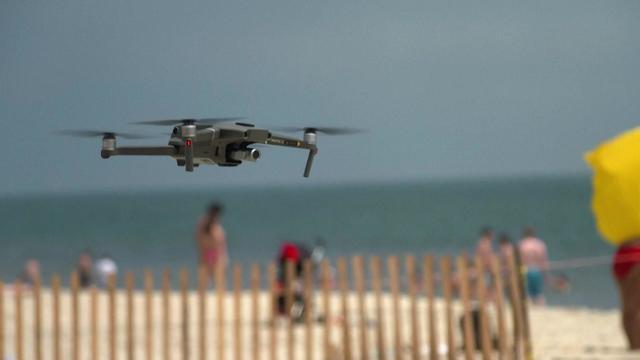 A drone hovers over a beach with beachgoers in the background. 
