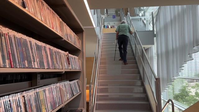 An individual using a cane walks up a flight of stairs inside the Hunters Point Library. 