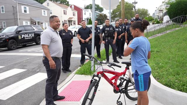 Nine Passaic Police members and the Passaic mayor talk to a 9-year-old boy standing with a new bicycle on a street corner. 