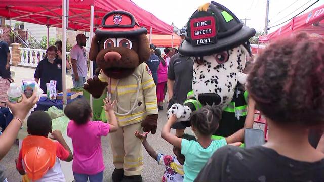 Two costumed mascots -- a brown dog dressed as a firefighter and a Dalmatian dressed as a firefighter -- interact with children at an FDNY block party. 