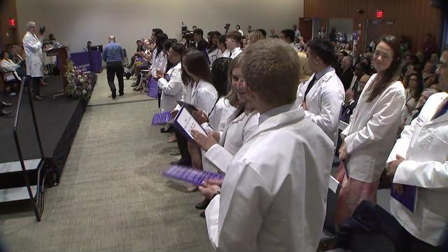 Medical students in white coats stand in the audience at an NYU Long Island School of Medicine ceremony. 