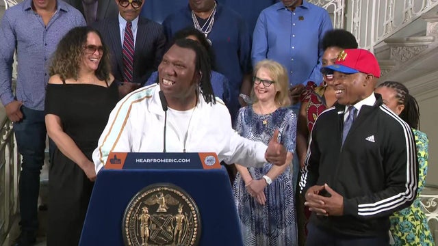 KRS-One stands behind a podium next to Mayor Eric Adams, who is wearing a tracksuit and baseball hat. 