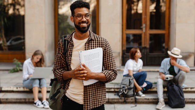 Happy young university student with book standing outdoors in front of campus. Back to university concept. 