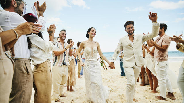Wide shot of bride and groom walking down aisle after wedding ceremony on tropical beach while friends and family celebrate 