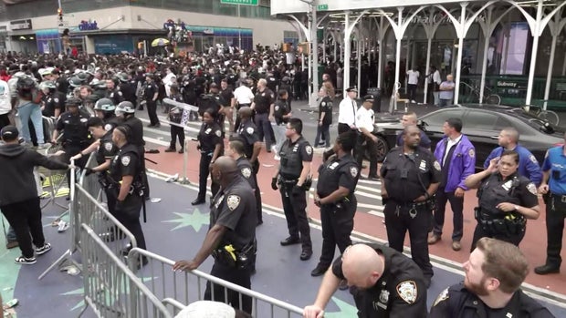 NYPD officers stand behind barriers set up near Union Square as hundreds of people crowd in the background.