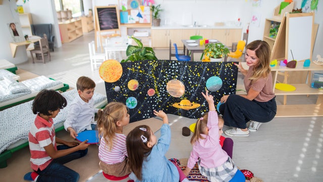 High angle view of pre school teacher showing poster with planets to children indoors in nursery, Montessori education. 