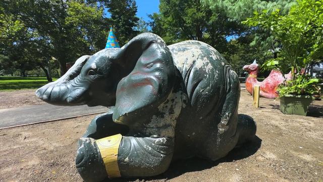 A weathered elephant statue sporting a paper gold watch and party hat. A weathered camel statue sporting a party hat and paper gold watch is in the background. 