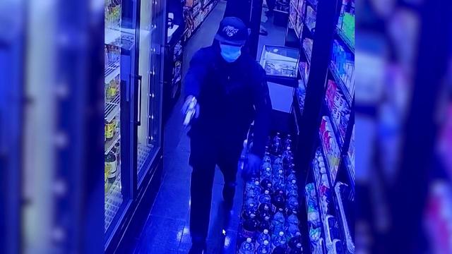 Surveillance video shows an individual wearing a mask, hoodie and hat pointing a gun down the aisle of a smoke shop. 