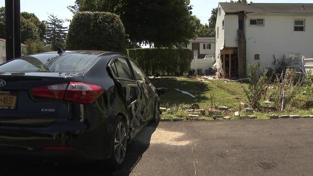 A sedan with severe damage to its passenger side sits parked. In the background, a home with damage to the exterior is seen. 