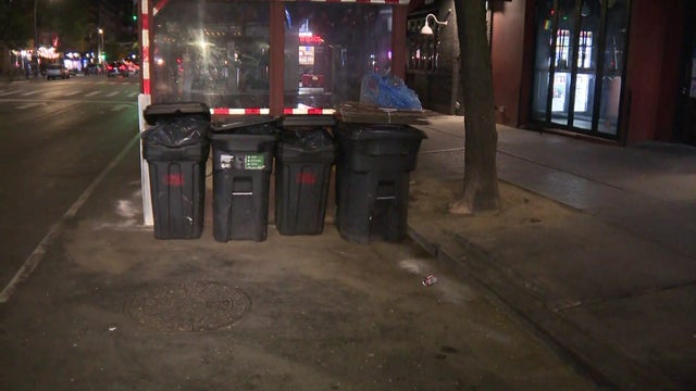 Four trash bins sit next to an outdoor dining structure on a New York City street. 