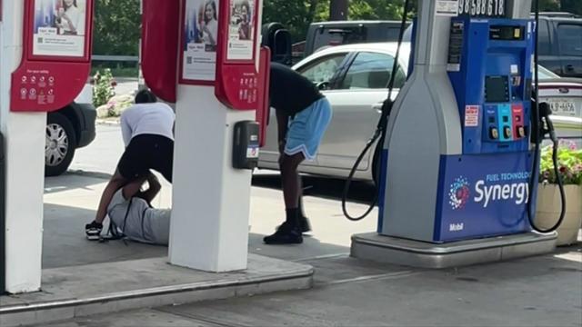 An individual is seen attacking a woman on the ground near gas station pumps while another individual films with a cellphone. 
