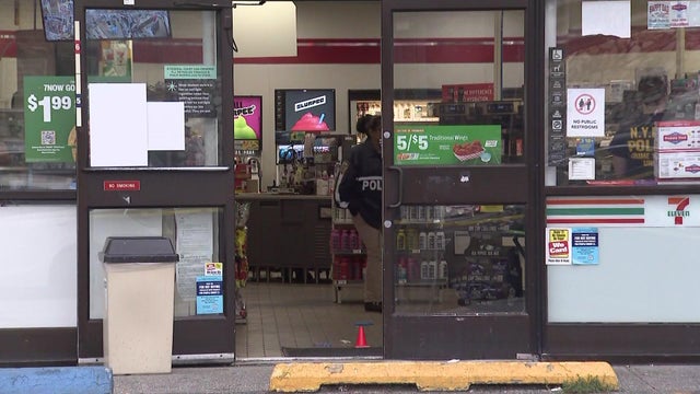 NYPD officers investigate inside a 7-Eleven. An orange cone marker can be seen on the floor in the doorway. 