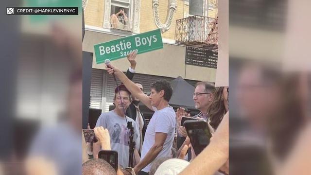 Members of the Beastie Boys stand on a stage as a street sign reading "Beastie Boys Square" is lifted into the air. 