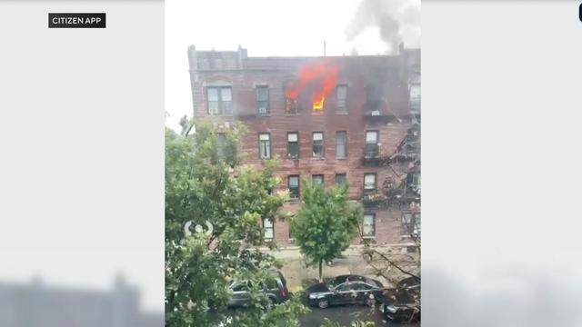Flames can be seen pouring from two windows on the top floor of an apartment building. 