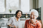 An elderly woman laughs beside a friendly young care assistant 