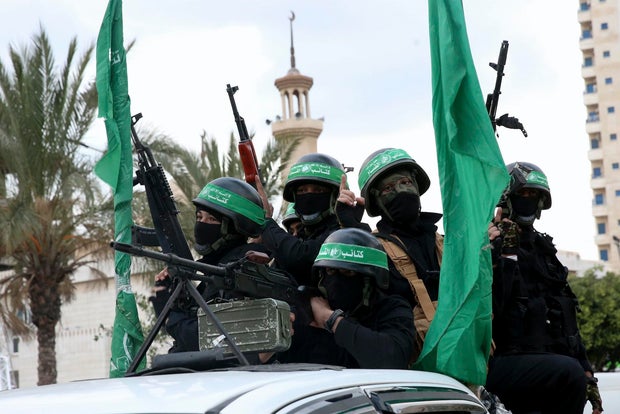 five fighters, armed with rifles and wearing helmets with green bands, look out from the bed of a truck
